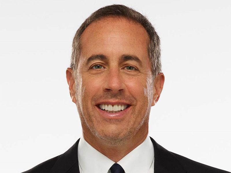 Smiling Jerry Seinfeld wearing white shirt and black coat